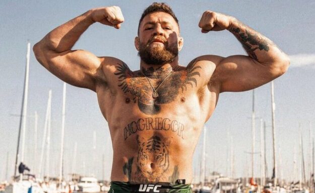 A man with a beard and tattoos strikes a pose against a backdrop of boats, exuding a cool, relaxed vibe.